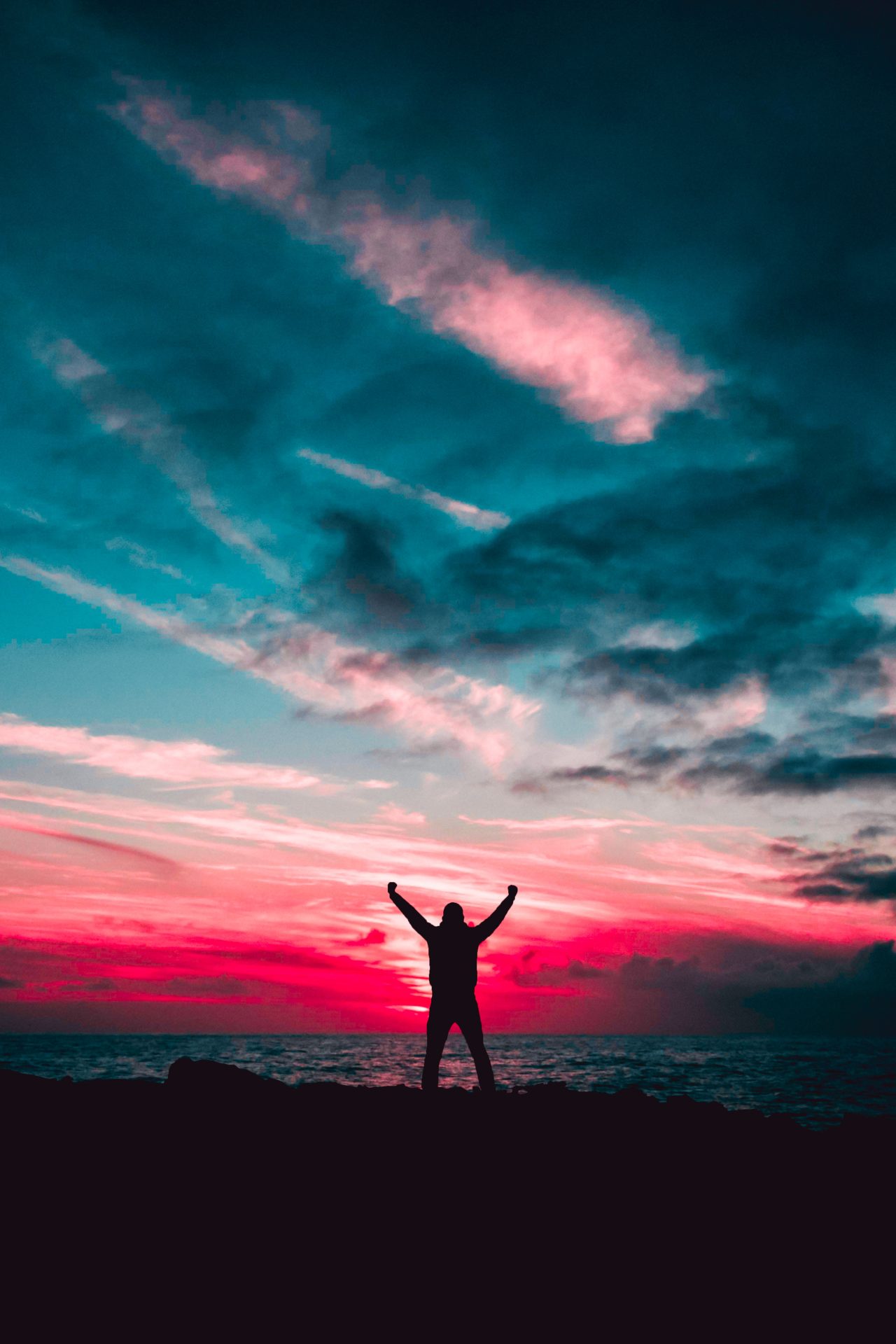 Person with arms raised as if in victory standing in front of a vibrant pink and blue sunrise or sunset over water.
